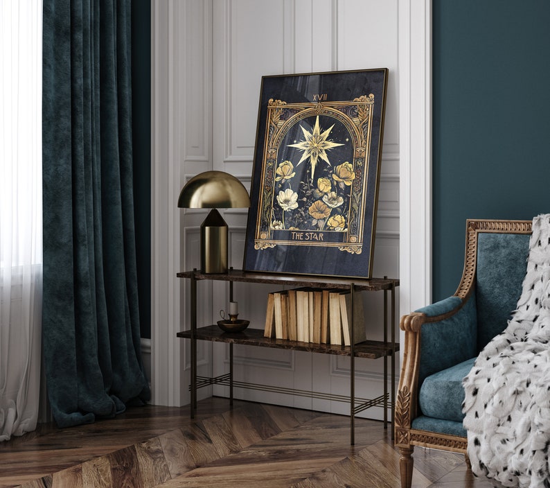 May include: A gold framed tarot card print with a dark blue background. The card depicts a star with yellow flowers and the text "XVII THE STAR". The print is hanging on a wall above a shelf with books and a lamp.