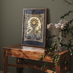 May include: A framed tarot card print with a gold and black design. The card depicts a sun with rays and sunflowers. The card is titled "The Sun" and numbered "XIX".