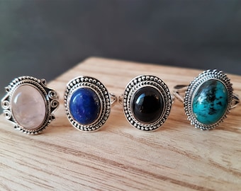 May include: Four silver rings with oval gemstones: rose quartz, lapis lazuli, black onyx, and turquoise. Each ring has a detailed silver setting with a beaded border. The rings are displayed on a light wooden surface.
