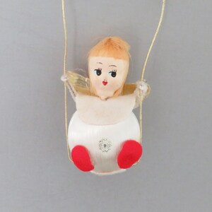 May include: A vintage Christmas ornament of a doll with blonde hair, wearing a white dress and red shoes, swinging on a gold string.