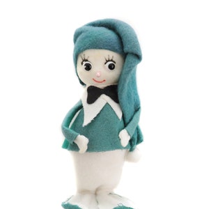 May include: A handmade felt doll with a teal-colored hat and sweater, a white body, and a black bow tie. The doll has a smiling face with rosy cheeks and long eyelashes. The doll's feet are teal and white, and it is standing upright.