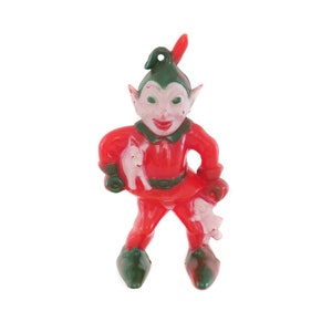 May include: A red plastic Christmas elf ornament with green pointed ears and hat. The elf is holding two small white plastic reindeer in its hands.