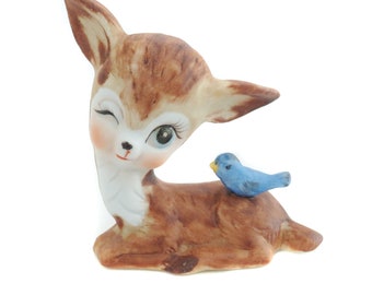 May include: A ceramic figurine of a brown and white deer with large eyes and a blue bird perched on its back. The deer is winking and the bird has a yellow beak.