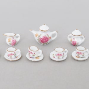 Op de afbeelding: Een miniatuur porseleinen theeservies met een theepot, suikerpot, melkkan en zes kopjes met schotels. Elk stuk is wit met een gouden rand en versierd met roze en rode bloemmotieven. Een charmant verzamelobject.