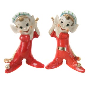 May include: Two vintage ceramic figurines of smiling elves in red suits and green and white striped hats. The elves have gold accents and are holding golf clubs. Each figurine has a silver ball on the foot.