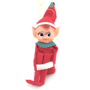 May include: A vintage Christmas elf figurine. The elf is dressed in a red felt outfit with a white belt and a red hat with a white brim. The elf has a plastic face with blue eyes, rosy cheeks, and a green collar. A yellow string is attached to the hat.