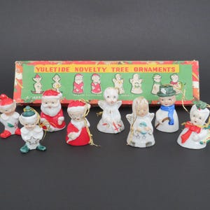 May include: A set of ten vintage porcelain Christmas tree decorations in a box. The decorations feature a variety of characters, including Father Christmas, angels, elves, and a snowman. The box is red and green with the words "Yuletide Novelty Tree Ornaments" printed on it.