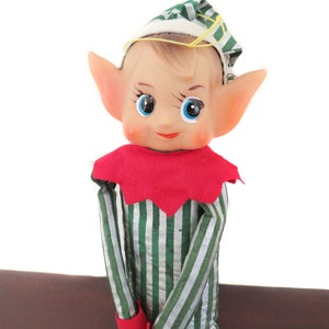 May include: A vintage elf doll with a painted face, large ears, and blue eyes. The doll wears a red collar, a green and white striped outfit, and a matching cap. The doll is a collectible item.