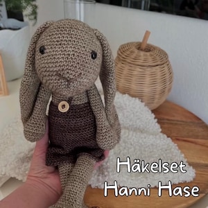 Könnte beinhalten: Handgemachte Häkelkaninchenpuppe in Brauntönen, mit braunen Shorts und einem Holzknopf. Das Kaninchen hat lange Ohren und schwarze Knopfaugen. Der Text "Häkelset Hanni Hase" ist im Bild zu sehen.