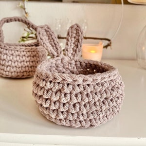 May include: Two handmade crochet baskets in a soft mauve color. One basket has bunny ears, and the other has a handle. The baskets are sitting on a white surface. A candle and a mirror are in the background.