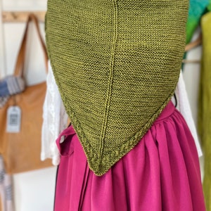 Puede incluir: Un chal triangular de punto verde con un borde decorativo. El chal se muestra en un maniquí que lleva una blusa blanca y una falda rosa.