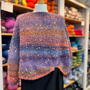 Puede incluir: Un suéter multicolor con lentejuelas sobre un maniquí. El suéter presenta tonos de morado, azul, marrón y rosa. La prenda tiene mangas largas y un cuello redondo. En el fondo se ven estantes con hilos de colores.