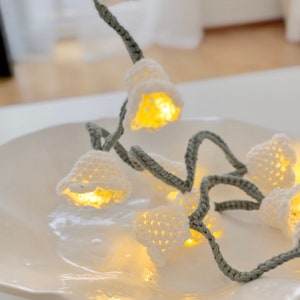 May include: A string of white crocheted flower lights with warm white LED bulbs. The lights are strung on a gray cord and are displayed on a white plate.