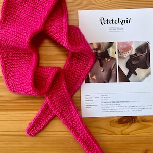 Könnte beinhalten: Ein leuchtend pinkfarbener, gestrickter Schal in spitz zulaufender Form, neben einer gedruckten PetiteKnit Sophie Scarf Anleitung. Der Schal hat eine kraus rechts gestrickte Textur und spitze Enden, während das Papierdokument Text und Bilder enthält.