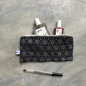 Op de afbeelding: Een zwarte en witte geometrisch gepatroneerde ritszak met een wit label met daarop "tine" aan de linkerkant. De zak is open en bevat een heldere glazen fles met een wit label met daarop "gitti natural spray" en een klein rood flesje met een wit label met daarop "g' 102". Een zwarte pen ligt op het grijze oppervlak.