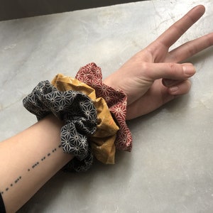 Könnte beinhalten: Drei Stoff-Haargummis in Schwarz, Gelb und Rot mit geometrischem Muster. Die Haargummis sind auf dem Arm einer Person gestapelt.