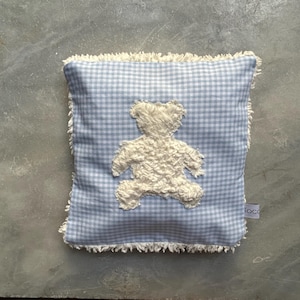 Op de afbeelding: Een blauw en wit geruit kussen met een witte, pluche teddybeer geborduurd op de voorkant. Het kussen heeft een witte franje rand.