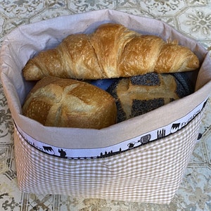 Op de afbeelding: Een beige stoffen broodmand met een ruitjespatroon en een zwart-witte rand. De mand bevat een croissant, een rond brood en een brood met maanzaad.