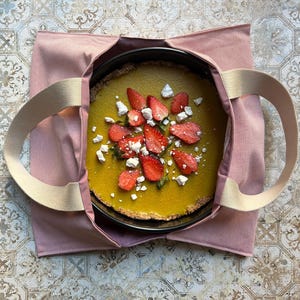 Könnte beinhalten: Eine rosa Stofftasche mit beigefarbenen Henkeln, in der eine runde Torte mit gelber Füllung liegt, die mit frischen Erdbeeren und weißem Baiser belegt ist.