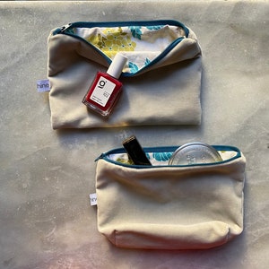 Op de afbeelding: Twee beige ritszakjes met turquoise bies. Het bovenste zakje is open en bevat een rode nagellak fles met de tekst "g 102" op het etiket. Het onderste zakje is open en bevat een ronde zilveren container en een zwarte make-up stick.