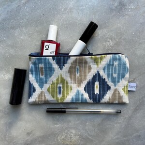 Könnte beinhalten: Eine Kosmetiktasche mit Reißverschluss und einem Muster aus blauen, grünen und braunen Rauten, die roten Nagellack, einen schwarzen Marker und einen schwarzen Lippenstift enthält. Ein durchsichtiger Stift liegt neben der Tasche.