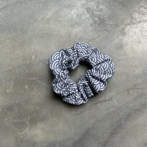 Könnte beinhalten: Ein Haargummi mit einem sich wiederholenden Wellenmuster in Weiß und Grau. Der Stoff ist gerafft, wodurch eine strukturierte, kreisförmige Form entsteht. Dieses Haaraccessoire ist für Pferdeschwänze oder Duttfrisuren konzipiert.