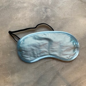 Könnte beinhalten: Eine hellblaue Schlafmaske aus Satin mit einem schwarzen Gummiband. Die Maske hat eine geschwungene Form, um sich den Augen anzupassen, und ist mit passendem hellblauem Satin eingefasst. Die Schlafmaske soll das Licht blockieren.