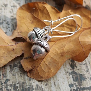 May include: Silver acorn earrings with a detailed design, hanging from silver hooks. The earrings are displayed on a brown leaf.
