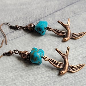 May include: A pair of turquoise and copper earrings featuring a small turquoise stone and a copper bird charm. The earrings are dangle style and have a hook closure.