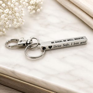 May include: A silver-toned metal bar keychain featuring a lobster clasp and a circular ring. The rectangular bar is engraved with the text BE GOOD, BE WELL, BEHAVE and Drive Safe, I love you! accompanied by a small black heart symbol.