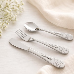 May include: A set of three silver-toned stainless steel utensils including a spoon, a fork, and a knife. Each handle is engraved with the name Anastasia and features a small embossed bear icon at the end of the handle with a smooth finish.
