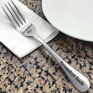 May include: A stainless steel fork with the words "Personalized Tines" and "Personalized Handle" engraved on it. The fork is resting on a white napkin next to a white plate, set on a speckled countertop.