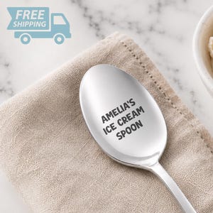 May include: A polished silver-toned metal spoon featuring the engraved black text AMELIA'S ICE CREAM SPOON centered on the bowl of the spoon. The spoon has a smooth, reflective surface and a classic rounded shape, resting on a textured beige fabric.