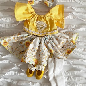 May include: A yellow short-sleeved top with a white lace heart and floral trim, a skirt with a yellow and white daisy print, a matching daisy print hair bow, a pair of yellow shoes with ruffled white trim, and white ribbed leggings.