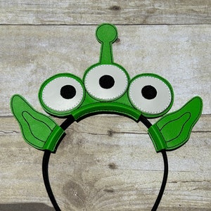 Könnte beinhalten: Grüner Alien-Haarreifen mit drei Augen und Ohren. Der Haarreifen ist aus Filz und hat ein schwarzes Gummiband.