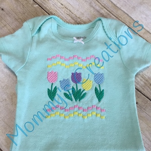 May include: A light blue baby onesie with a floral design. The onesie features an embroidered pattern of tulips in pink, purple, and yellow, with green stems and leaves. The text "Mommy's creations" is written on the onesie.