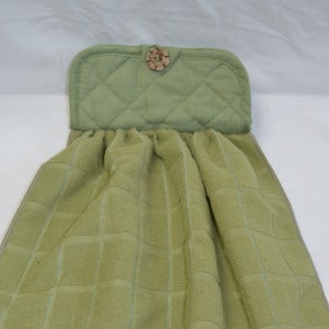 May include: A green kitchen towel with a quilted green fabric pocket and a wooden button. The towel has a checkered pattern.