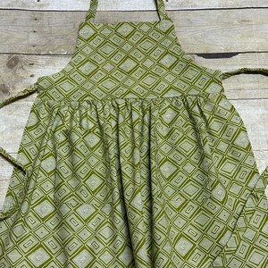 May include: A green apron with a white geometric pattern. The apron has adjustable straps and a gathered skirt.
