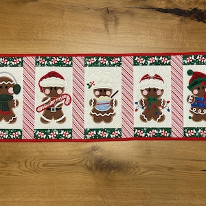 Puede incluir: Camino de mesa navideño rectangular con cinco diseños de hombre de jengibre. Cada hombre de jengibre está decorado de forma única con accesorios como orejeras, gorro de chef y gorro de Papá Noel. El camino de mesa tiene un borde rojo y detalles de rayas de bastón de caramelo.