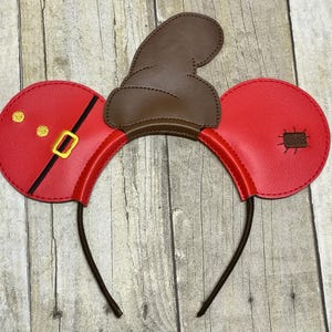 May include: Red and brown Mickey Mouse ears headband with a brown felt hat on top. The ears have gold accents and stitching.