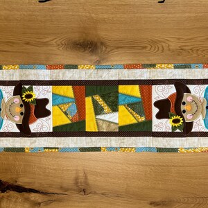 Op de afbeelding: Een rechthoekige, gewatteerde tafelloper met een patchwork-ontwerp. Het heeft een centraal paneel met kleurrijke geometrische vormen en twee vogelverschrikker-appliqués. De vogelverschrikkers hebben bruine hoeden en zonnebloemaccenten. De rand is veelkleurig.
