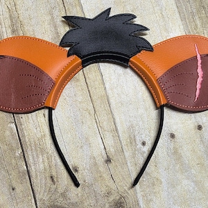 May include: A pair of orange and brown mouse ears with black hair on top. The ears are made of felt and have a black headband.