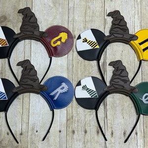 Könnte beinhalten: Vier Paar schwarze Mäuseohren mit verschiedenen farbigen Stoffpatches. Jedes Paar hat einen braunen Filz-Sortierschuh obenauf. Die Stoffpatches zeigen eine Krawatte, eine Schlange, ein Gryffindor-Wappen und ein Hufflepuff-Wappen.