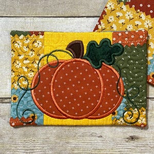 Puede incluir: Un posavasos rectangular acolchado con una gran calabaza naranja con una hoja verde y un tallo marrón. La calabaza está centrada en un fondo amarillo, rodeada de tela de retazos en colores otoñales. Un segundo posavasos es visible.