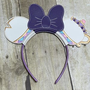Puede incluir: Diadema con lazo morado y orejas de taza de té blanca. Las tazas de té tienen ribetes dorados y un patrón rosa, azul y morado.