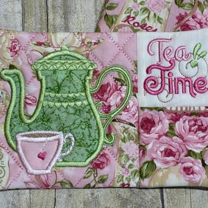 May include: Pink quilted tea cozy with a green teapot and teacup embroidery. The words "Tea Time" are embroidered in pink. The background features a floral pattern with roses.