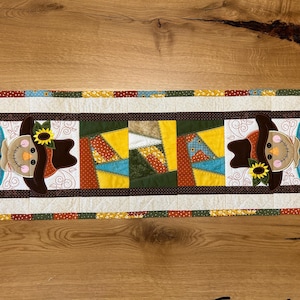 Op de afbeelding: Een rechthoekige, gewatteerde tafelloper met een patchwork-ontwerp met een herfstthema. De loper heeft een middenpaneel met een patchwork-ontwerp en twee vogelverschrikkerfiguren met zonnebloemen. De rand is crèmekleurig met een stippenpatroon.