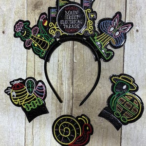 May include: Black headband with Main Street Electrical Parade design. Features a train, a butterfly, a bee, a snail, and a turtle. The text "Main Street Electrical Parade" is visible on the train. The designs are outlined in colourful lights.