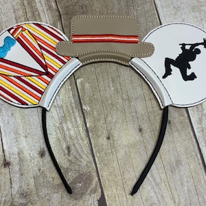 May include: Novelty mouse ears headband with a tan hat and two round ears. One ear features a colorful striped design with a blue bow, the other a black silhouette of a person. The headband is black.