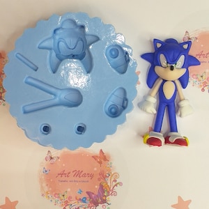 Può includere: Stampo in silicone blu con un design di Sonic the Hedgehog, che include la sua testa, le sue scarpe e un anello. Lo stampo è su una superficie bianca con stelle rosa e un design floreale rosa e bianco con farfalle.